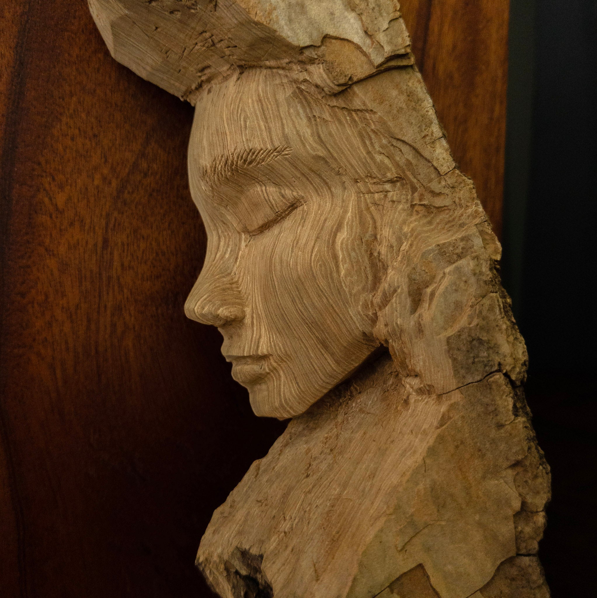 Close up of a hand carved sculpture of a young woman's face against a mahogany background
